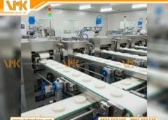 Một chiếc máy làm bánh gạo công nghiệp đang hoạt động, có nhiều đường băng chuyền với bánh gạo được tạo hình đều đặn, cho thấy quy trình sản xuất tự động và hiệu quả. Nền là không gian xưởng sản xuất hiện đại, sạch sẽ.